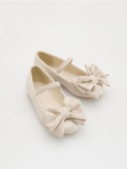 Lakierowane baleriny z kokardą - kremowy. Białe baleriny dziewczęce Reserved, bez wzorów, z lakierowanej skóry, bez obcasa, bez zapięcia. W wyprzedaży za 49.99 zł.